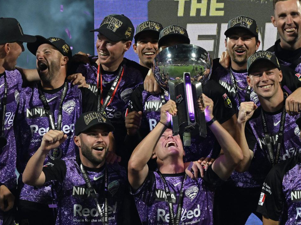 obart Hurricanes celebrate with 'NINJA' and 'Red' branding, wearing champion caps, holding the BBL trophy, laughing and cheering.
