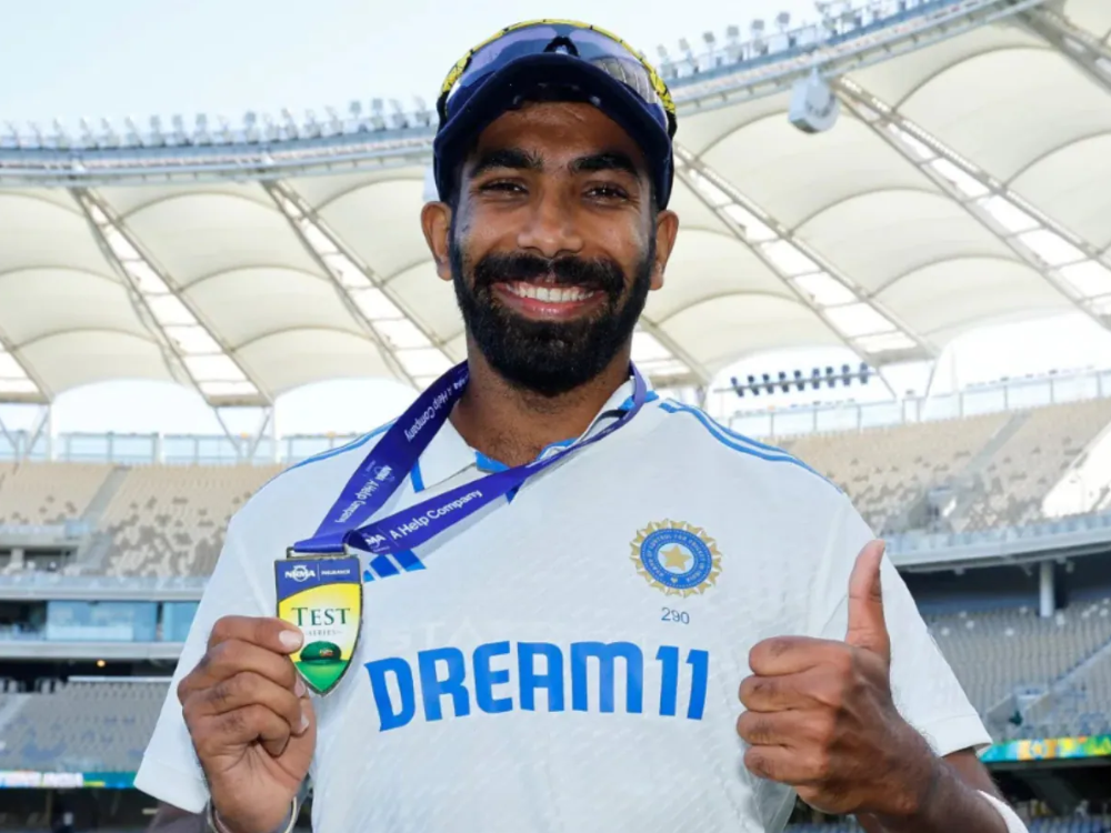 Jasprit Bumrah in a white India jersey with 'DREAM11' sponsor, sunglasses, blue cap, smiling, holding a Test cricket medal at a stadium.