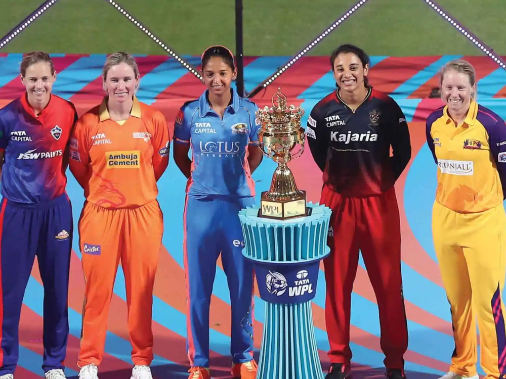 Five WPL captains in team jerseys stand with the WPL trophy on a blue pedestal, set against a vibrant backdrop.